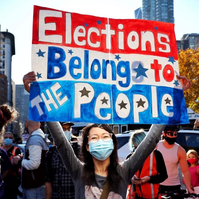 A photo of a protestor holding a sign reading "Elections belong to the people."